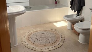Un baño con bañera, dos lavabos, un bidé, inodoro y un tapete tejido, además de un tragaluz.