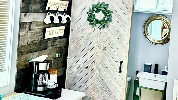 A rustic kitchen with a wooden herringbone wall, a coffee station, and a door with a wreath. - Peaceful Charming Tiny Home (Kyle)