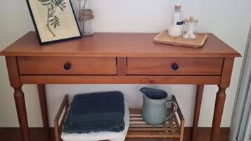 Een houten bureau met twee lades, een lijst met een botanische afbeelding, een kleine vaas met gedroogde bloemen, een zeepdispenser en een karaf op een houten plank eronder.