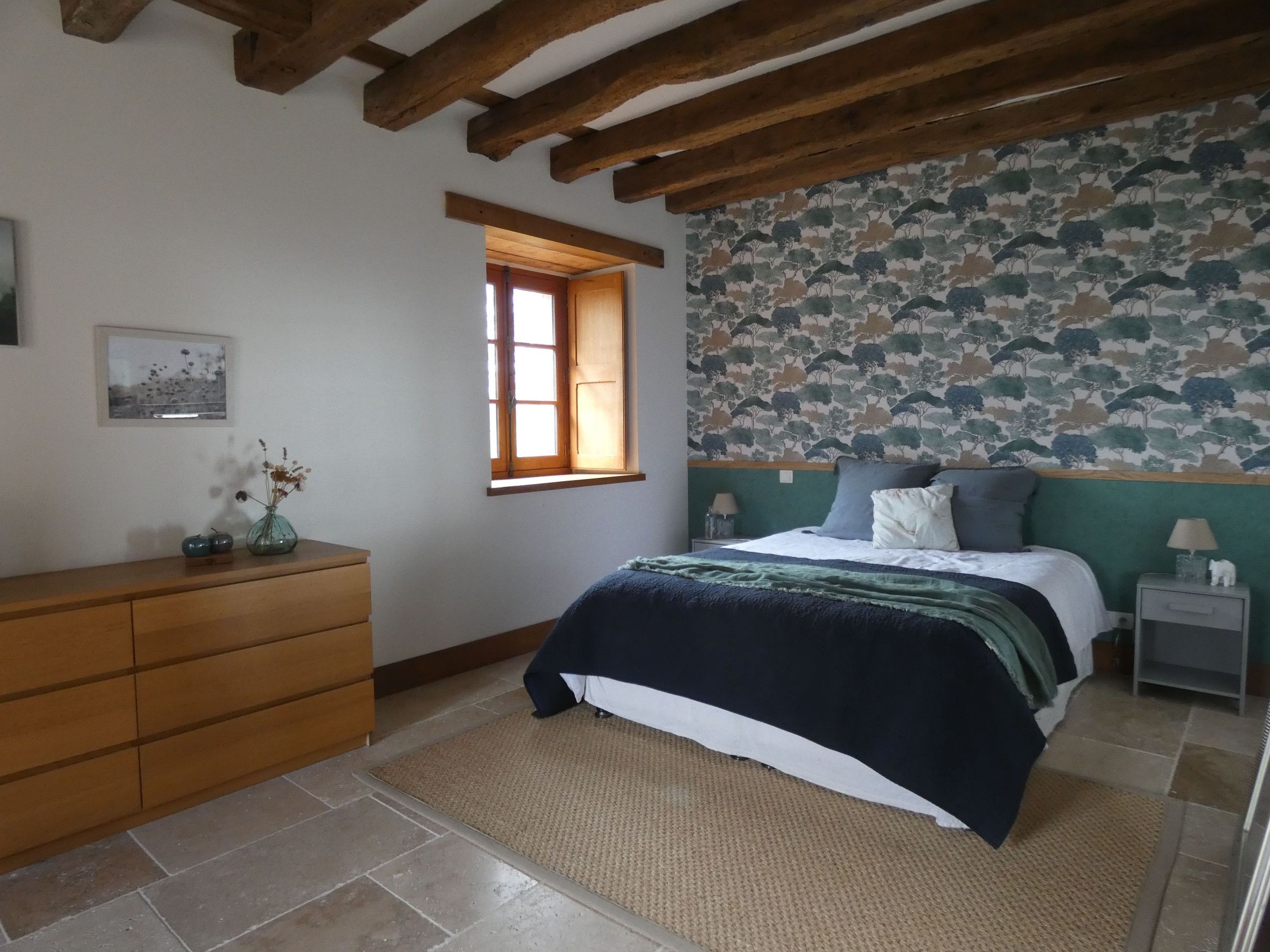 A bedroom with a bed, wooden dresser, a window, and a patterned wallpaper.