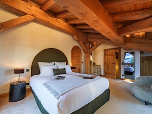 A bedroom with a large bed, wooden beams, a dresser, and a fluffy chair.