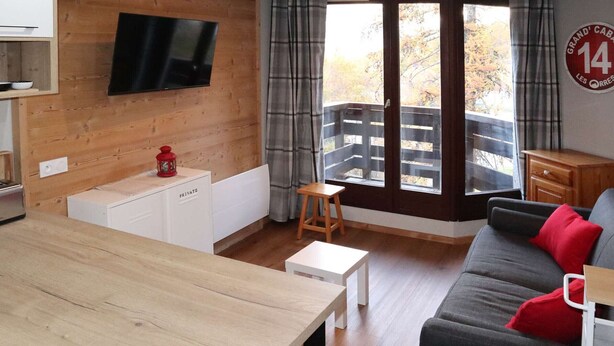 A modern living room with a wooden floor, a grey sofa with red cushions, a wooden dining table, a small white cabinet labeled 'PRIVATE', a television mounted on the wall, and a large window with curtains. - Renovated studio for 5, near lifts, balcony (Les Orres)