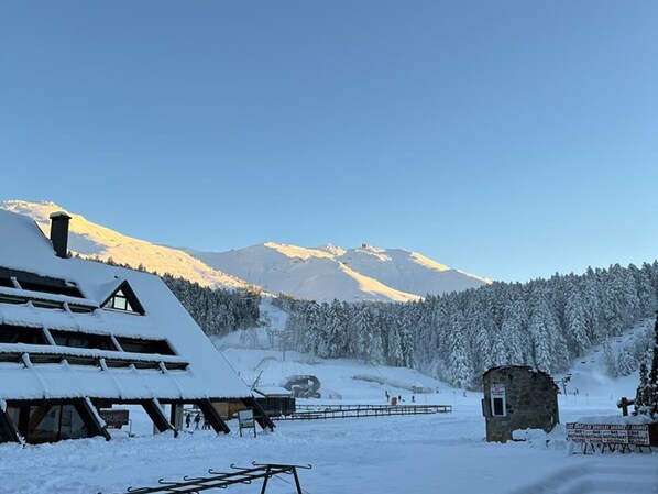 A snow-covered building with a sloped roof, a wooden deck, and a mountain backdrop.