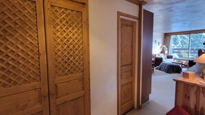 Een houten interieur met een verstelbare deur, uitzicht op een kamer met een bed en een raam, en een klein bureau met een lamp.