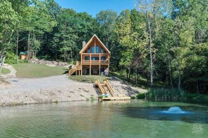 A wooden cabin with a balcony, situated by a lake surrounded by trees. - Lakeside Log Cabin | Hot Tub • Arcade • Views (West Lafayette)