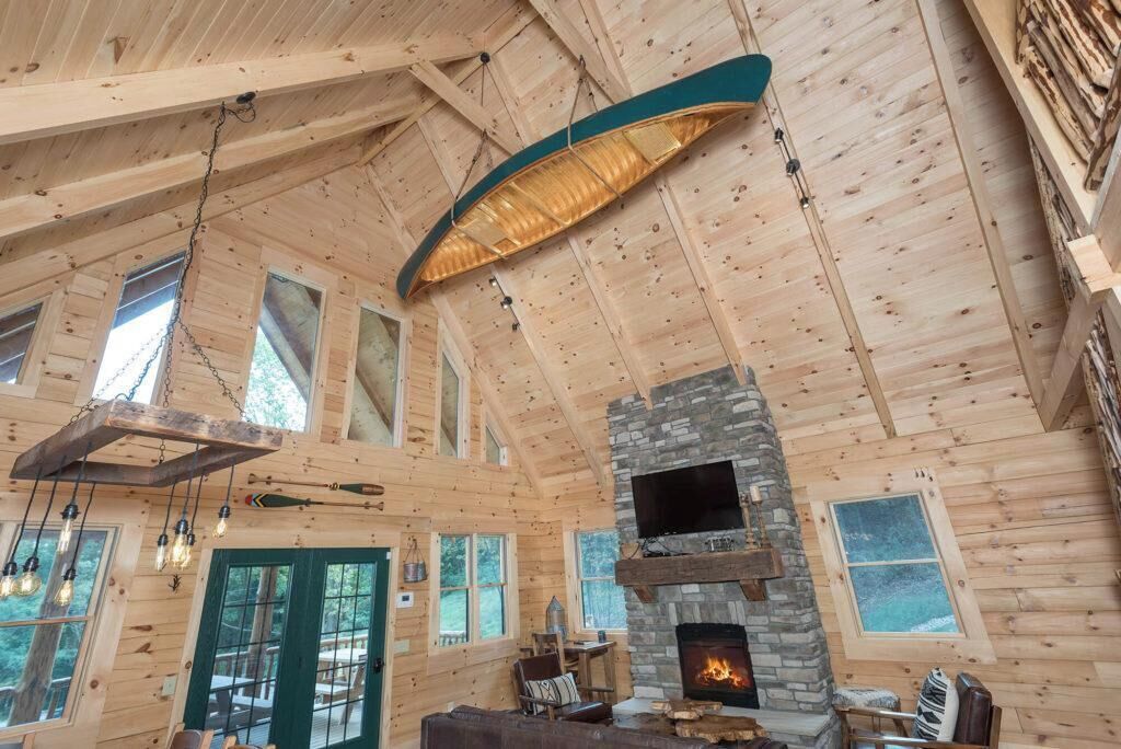 Interior de una cabaña de troncos con chimenea de piedra, vigas de madera y una lámpara colgante.