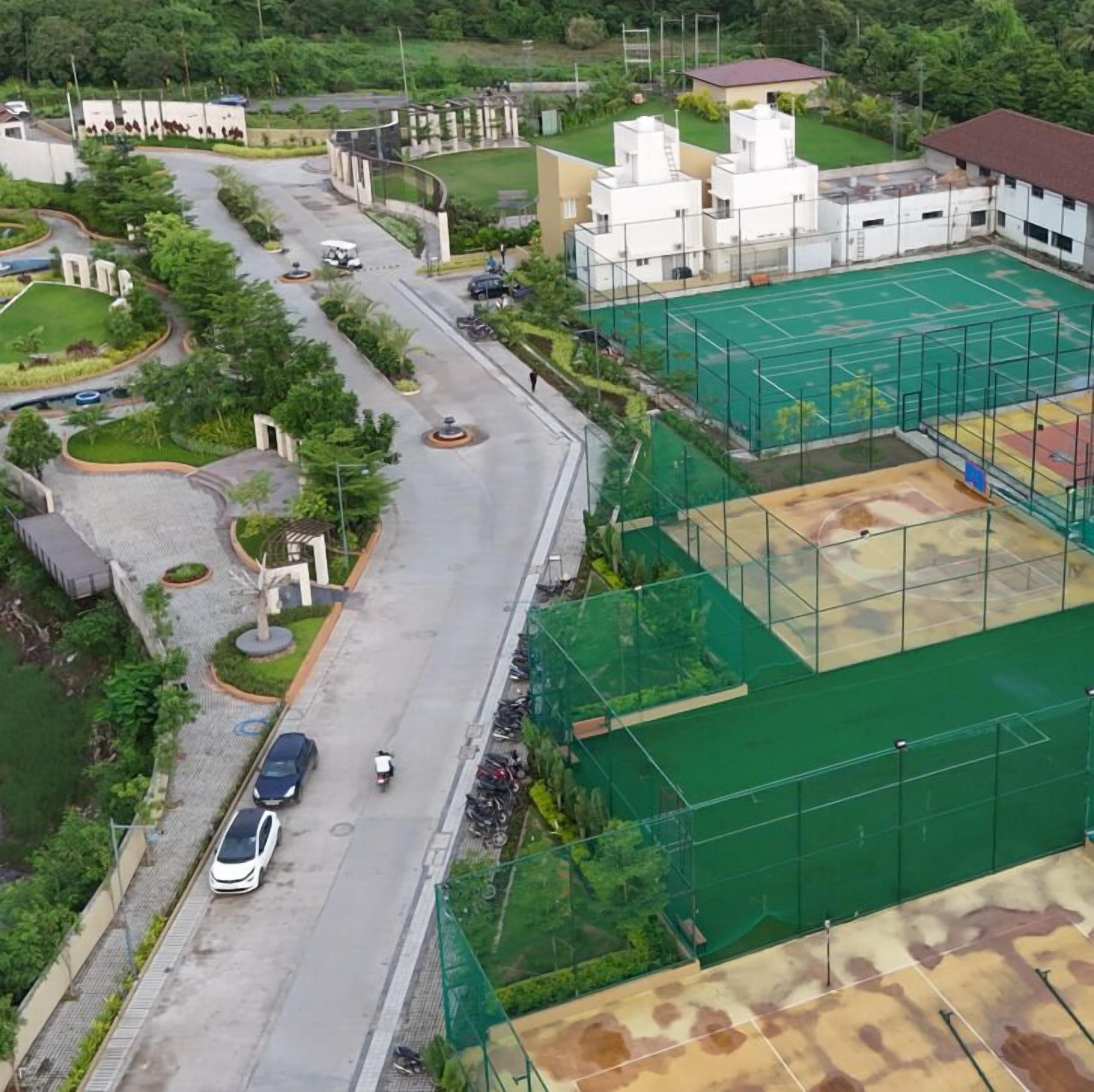 Vue aérienne d’un quartier résidentiel avec un court de tennis, une route où circulent des véhicules et des espaces verts environnants.