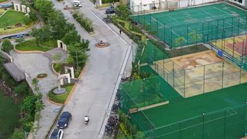 Vue aérienne d’un quartier résidentiel avec un court de tennis, une route où circulent des véhicules et des espaces verts environnants.