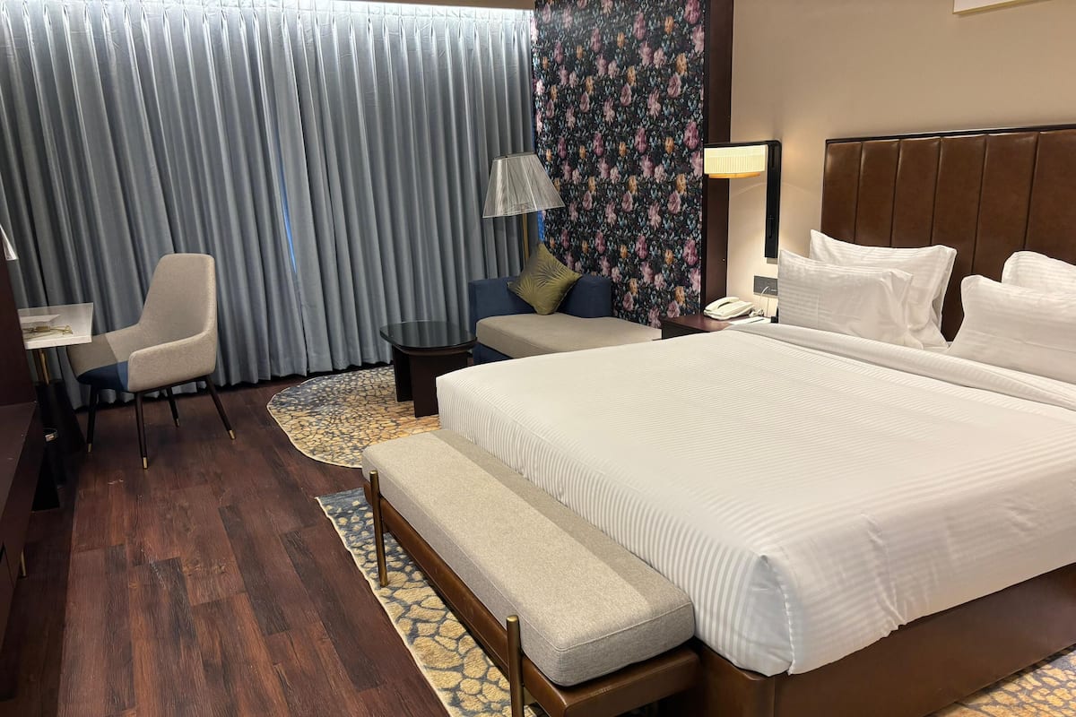 A hotel room with a large bed, a desk with a chair, a small table, and a lamp.