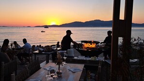 Un restaurant en bord de mer avec une terrasse, un foyer extérieur et une vue sur le coucher de soleil sur l’eau.