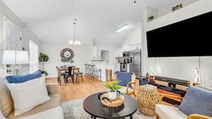 Un salon moderne avec un canapé, une table basse et des fauteuils. On y trouve également un coin repas avec une table et des chaises. Une cuisine équipée est visible au fond.