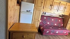 A compact kitchen with wooden cabinets, a white refrigerator, and a bed with a patterned blanket.