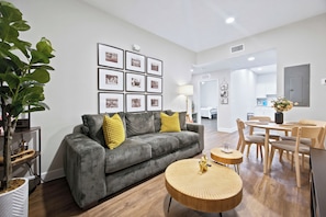 Una sala de estar moderna con un sofá gris, cojines amarillos, una mesa de centro redonda de madera y una zona de comedor con una mesa y sillas de madera.