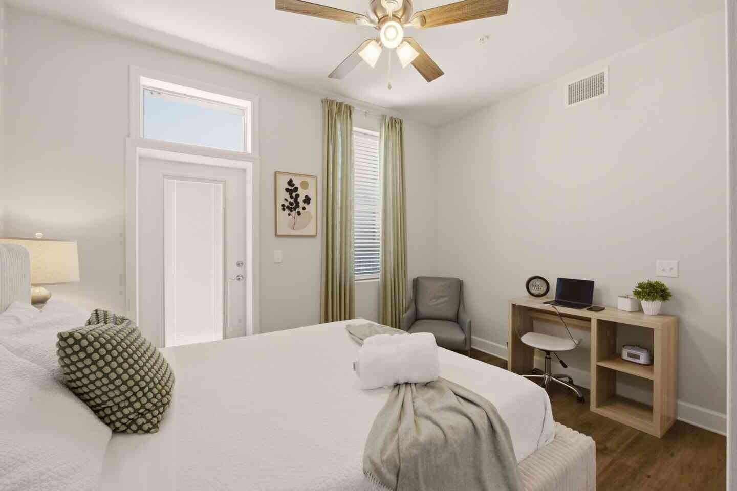 Habitación de hotel con cama, escritorio, silla, ventilador de techo y ventana con cortinas.