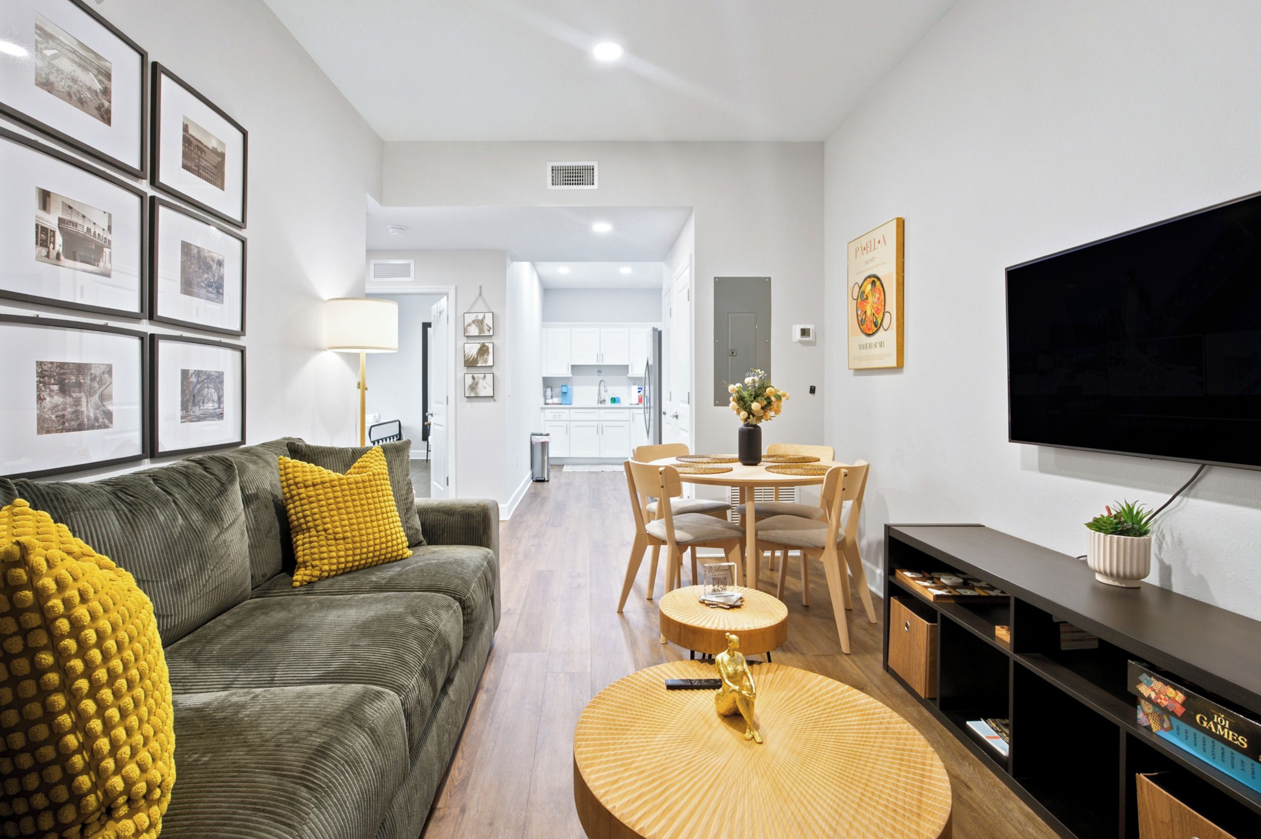 A modern living room with a sofa, coffee table, and a TV. There is a dining area with chairs and a table. The room has framed pictures on the wall and a kitchen in the background.