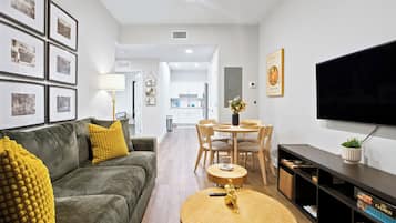 A modern living room with a sofa, coffee table, and a TV. There is a dining area with chairs and a table. The room has framed pictures on the wall and a kitchen in the background.