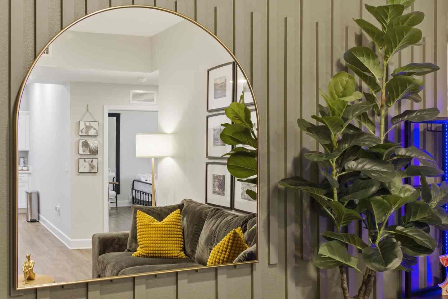 A living room with a large mirror, a sofa with yellow cushions, a potted plant, and framed pictures on the wall.