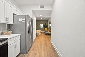 Una cocina moderna con muebles blancos, un refrigerador de acero inoxidable y una zona de comedor con sofá gris y mesa de madera.