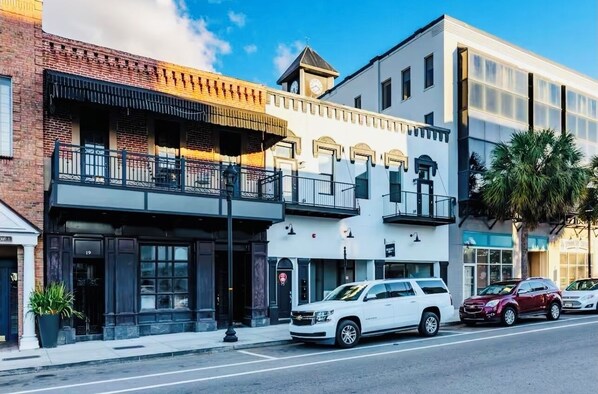 A street view with a two-story building featuring a balcony, a white car parked in front, and a palm tree. - Ocala Downtown Square Apartment (Ocala)