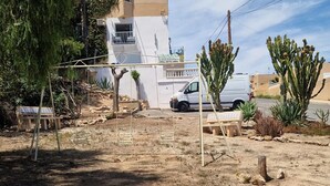 Paisaje desértico con una camioneta blanca estacionada cerca de un edificio con balcón. Se ven cactus y un banco de madera en primer plano.