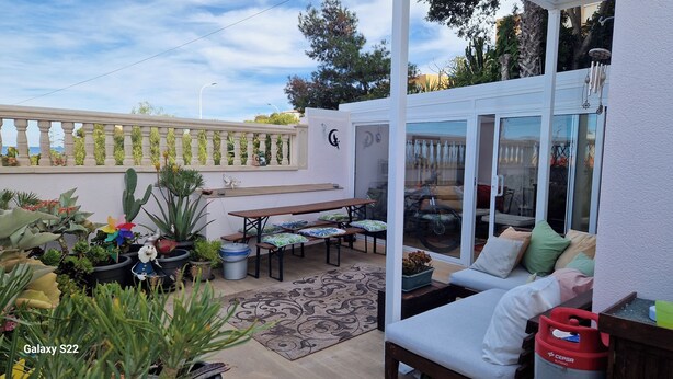 Un patio con mesa de comedor, bancos y sofá. Hay macetas con plantas y un molinillo decorativo.