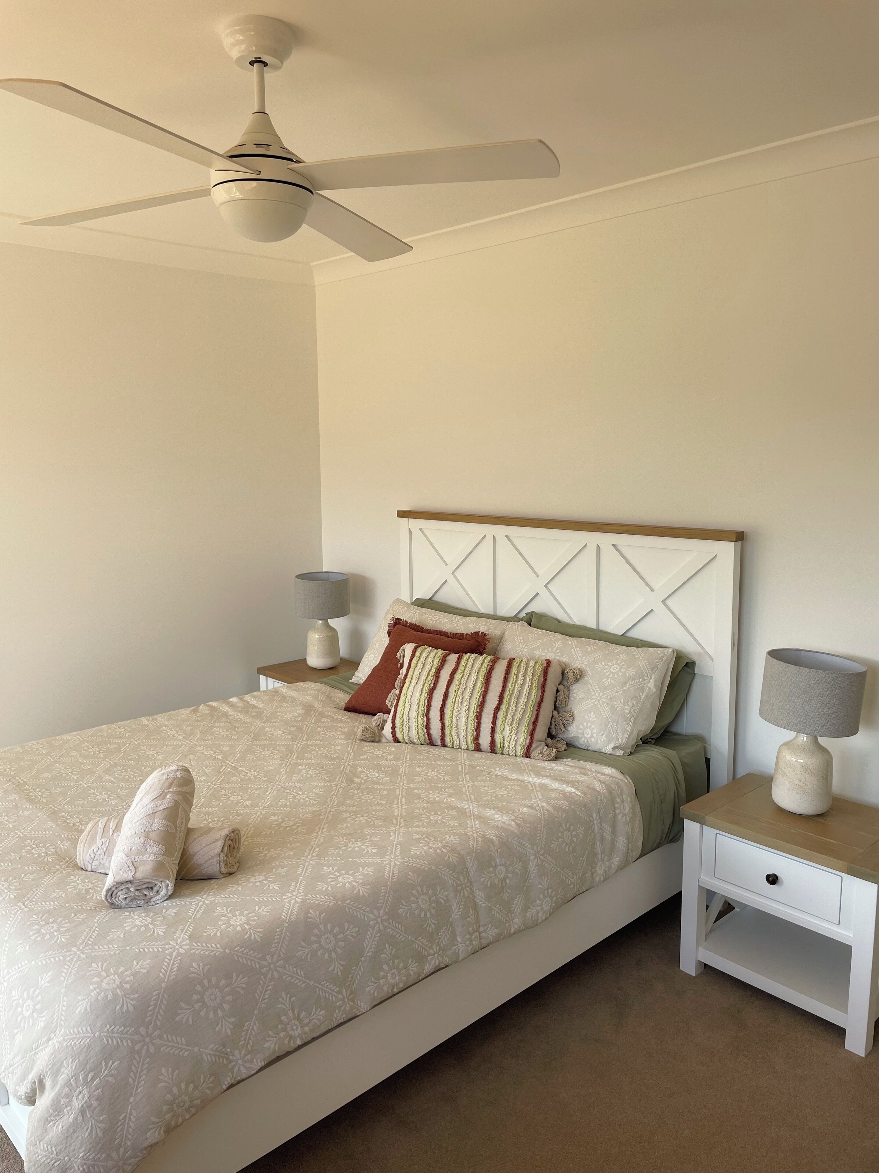 Un lit bien fait, avec une tête de lit blanche et un seul oreiller, une table de chevet avec une lampe et un ventilateur de plafond.