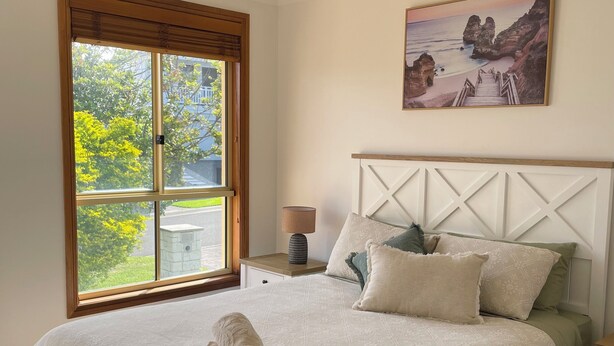 A bedroom with a large bed, a window with a view of greenery, a painting of a beach, and a bedside table with a lamp.