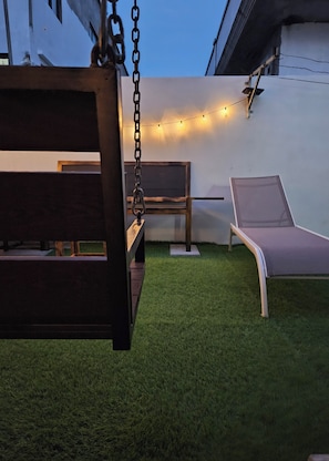 Une terrasse avec une balançoire, deux fauteuils de jardin et une guirlande lumineuse accrochée à un mur blanc.