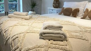 A neatly made bed with a beige textured blanket and pillows, a small bedside table with a vase, and a window with a view of greenery. - Luxury Family Home Central Bournemouth Sleeps 6 (Bournemouth)