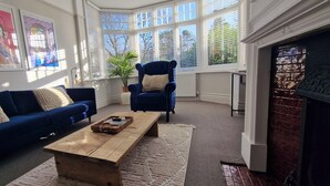 A living room with a blue sofa, a wooden coffee table, and a blue armchair. There are framed artworks on the wall, a potted plant, and a brick fireplace. - Luxury Family Home Central Bournemouth Sleeps 6 (Bournemouth)