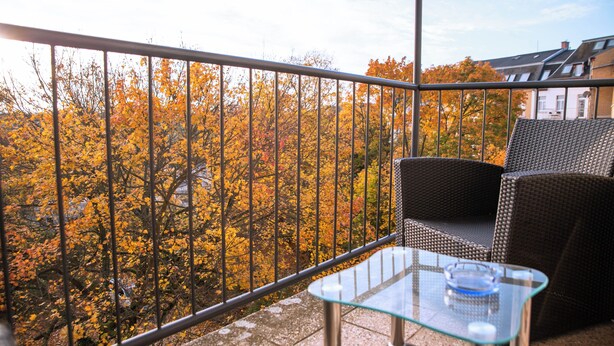 Un balcon doté d’une table en verre et de meubles en osier, offrant une vue sur les arbres aux couleurs automnales.