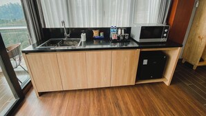 A modern kitchen with a black countertop, wooden cabinets, a stainless steel sink, a microwave, and a view of the outdoors through sheer curtains. - Damar Langit Resort (Cisarua)