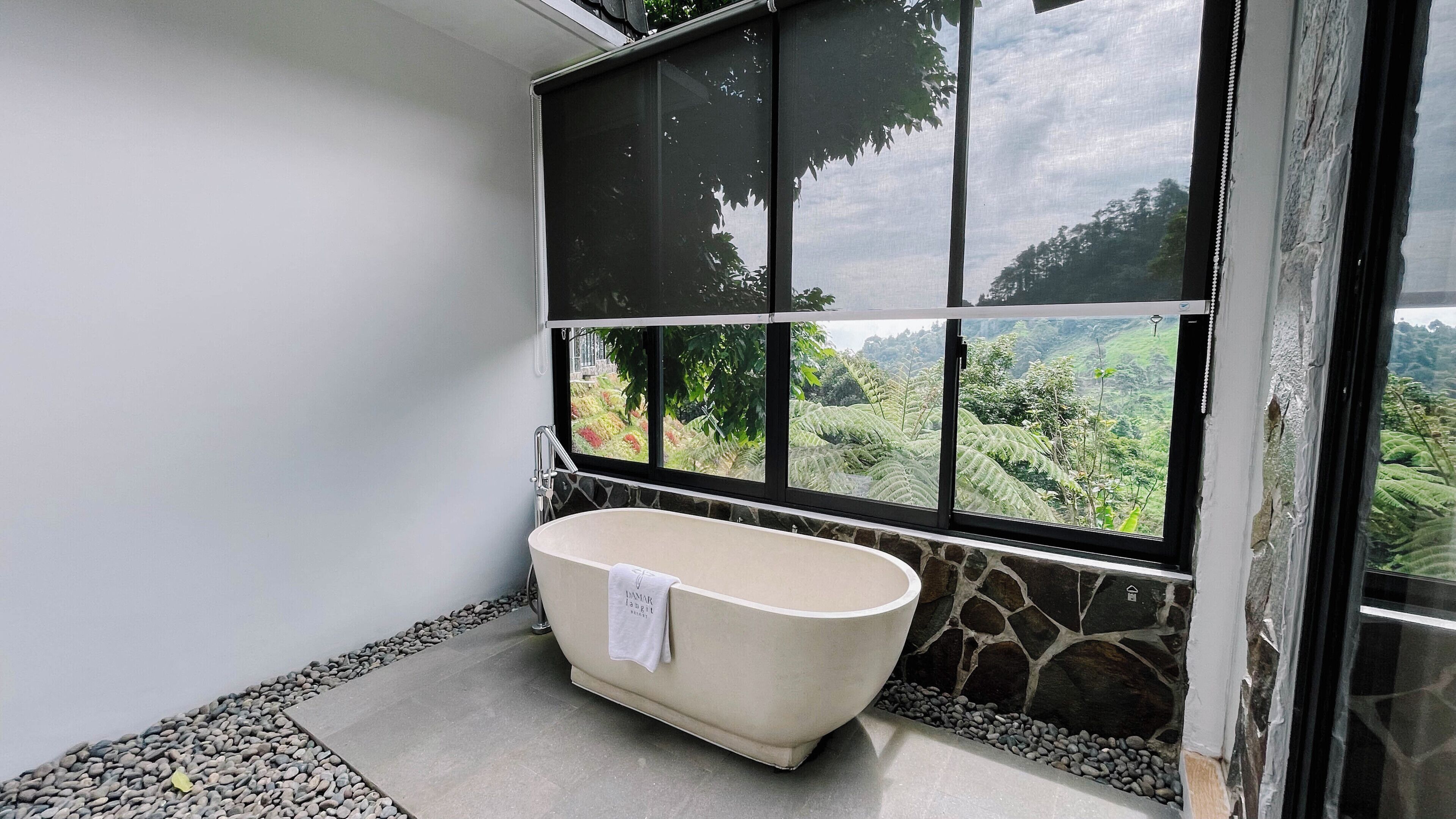 A bathroom with a freestanding bathtub, a stone wall, and a view of a lush landscape through large windows.