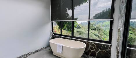 A bathroom with a freestanding bathtub, a stone wall, and a view of a lush landscape through large windows.