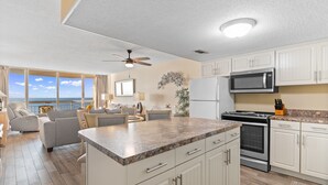 A modern kitchen with white cabinets, a granite countertop, and stainless steel appliances. There is a dining area with a view of the ocean. - Garden City Beach Oceanfront Condo 508 | Rising Tides Properties Rental (Murrells Inlet)