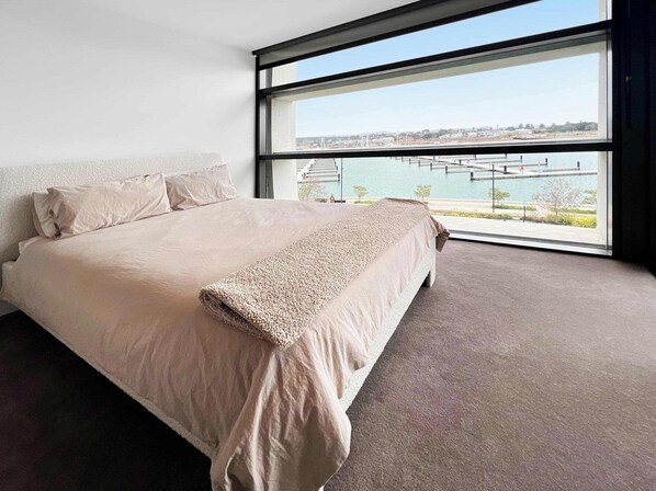 Una habitación de hotel moderna con una cama grande, vistas a un puerto deportivo y un ventanal amplio.