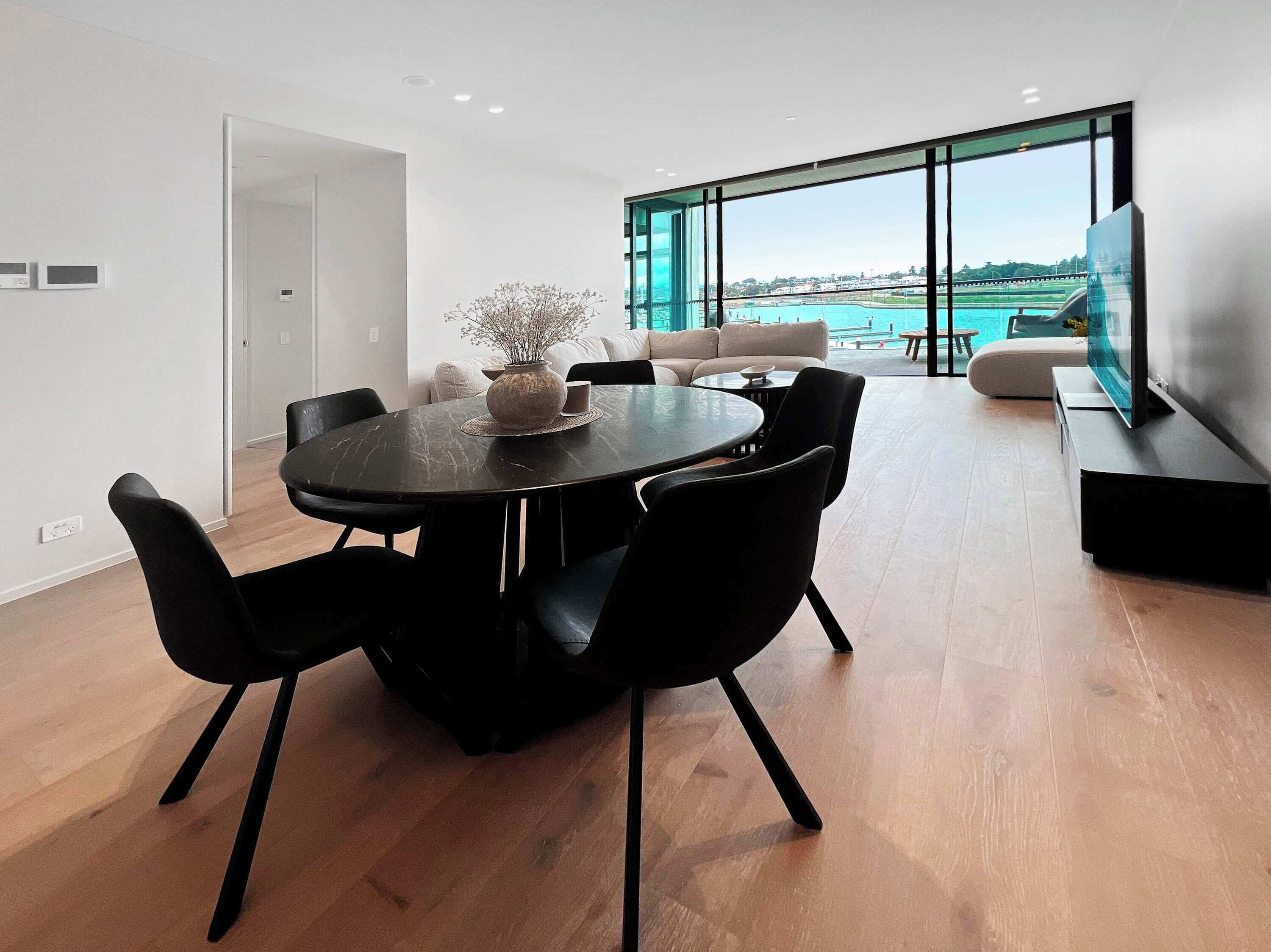 A modern dining area with a round table, black chairs, and a vase of flowers. There is a large window with a view of a body of water and buildings.