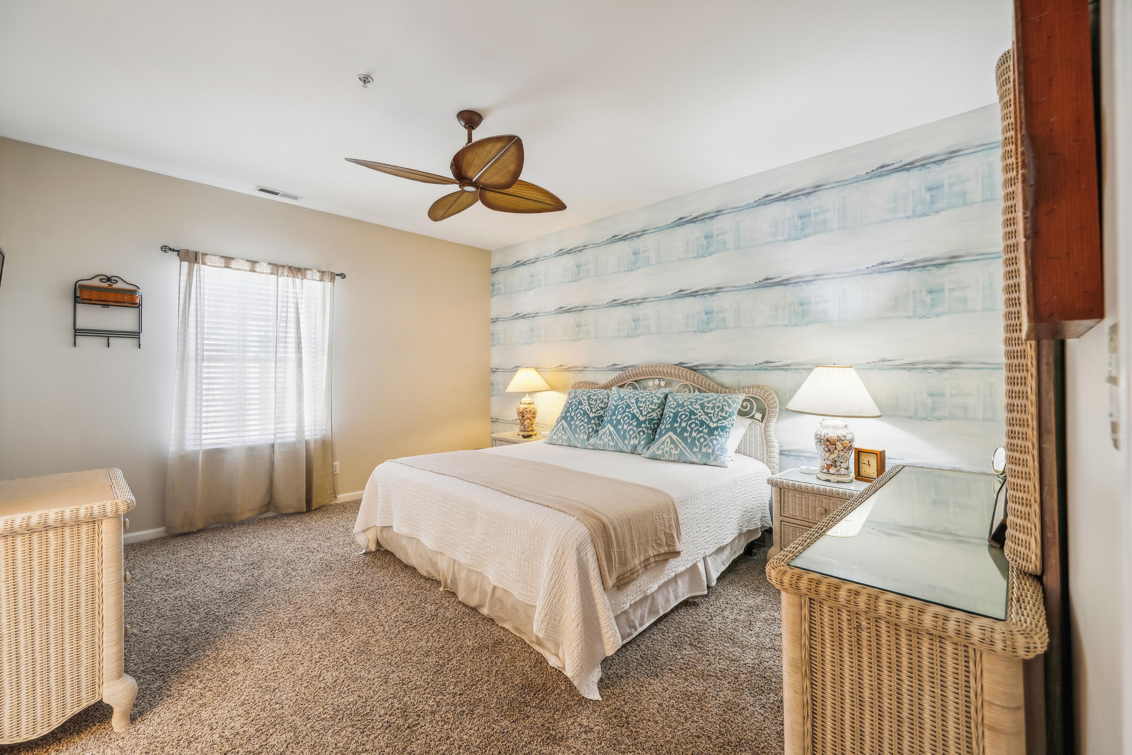 A bedroom with a bed, two bedside tables, a ceiling fan, a window with curtains, and a wall with a striped pattern.