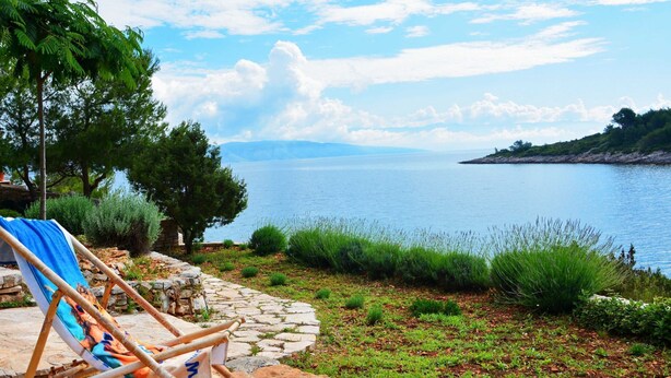 A stone pathway leading to a beach with a chair and a blanket, overlooking a calm sea and distant mountains.