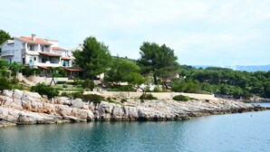 A coastal property with a stone retaining wall, a clear view of the sea, and a multi-story building with balconies.