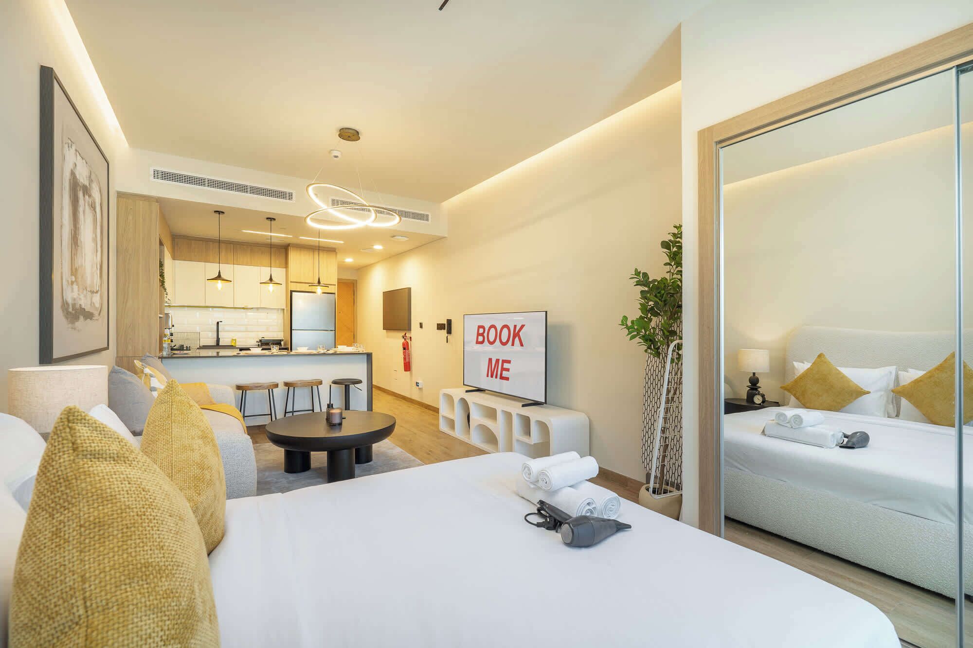 A hotel room with a bed, a desk with a chair, a small kitchen area, and a 'BOOK ME' sign.
