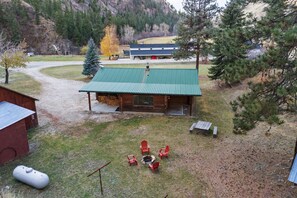 A log cabin with a green roof, surrounded by a grassy yard with red Adirondack chairs and a fire pit.