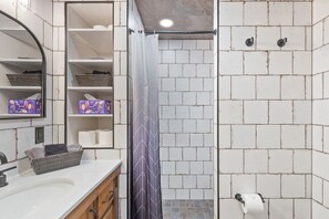 A bathroom with white tiled walls, a shower area with a curtain, a sink with a mirror, and shelves with baskets and towels. - Sapphire Ridge Cabin (Hamilton)