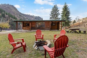A cabin with a fire pit and red Adirondack chairs in a grassy area with a mountain backdrop.