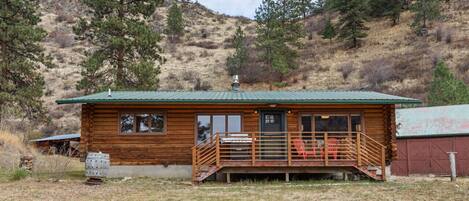 A wooden cabin with a green roof, a porch with a railing, and a small outdoor seating area.