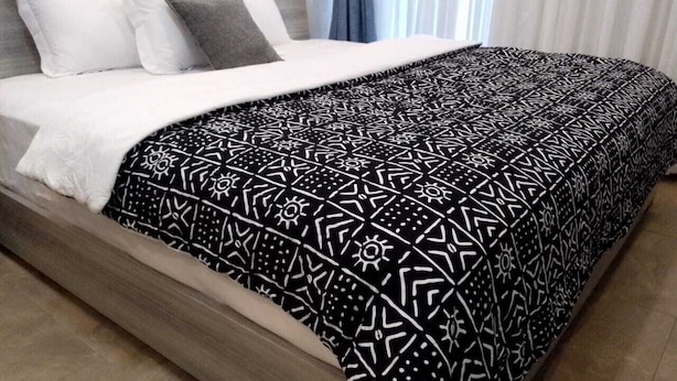 A bed with a patterned bedspread, a white pillow, and a grey pillow. - Hôtel-228 (Lomé)