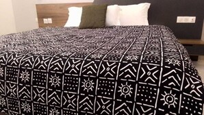 A neatly made bed with a patterned bedspread and two pillows, a wooden headboard, and a bedside table with a lamp. - Hôtel-228 (Lomé)