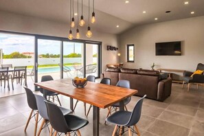A modern living room with a wooden dining table, black chairs, a brown leather sofa, and a TV mounted on the wall. - Villa Esti (Loborika)