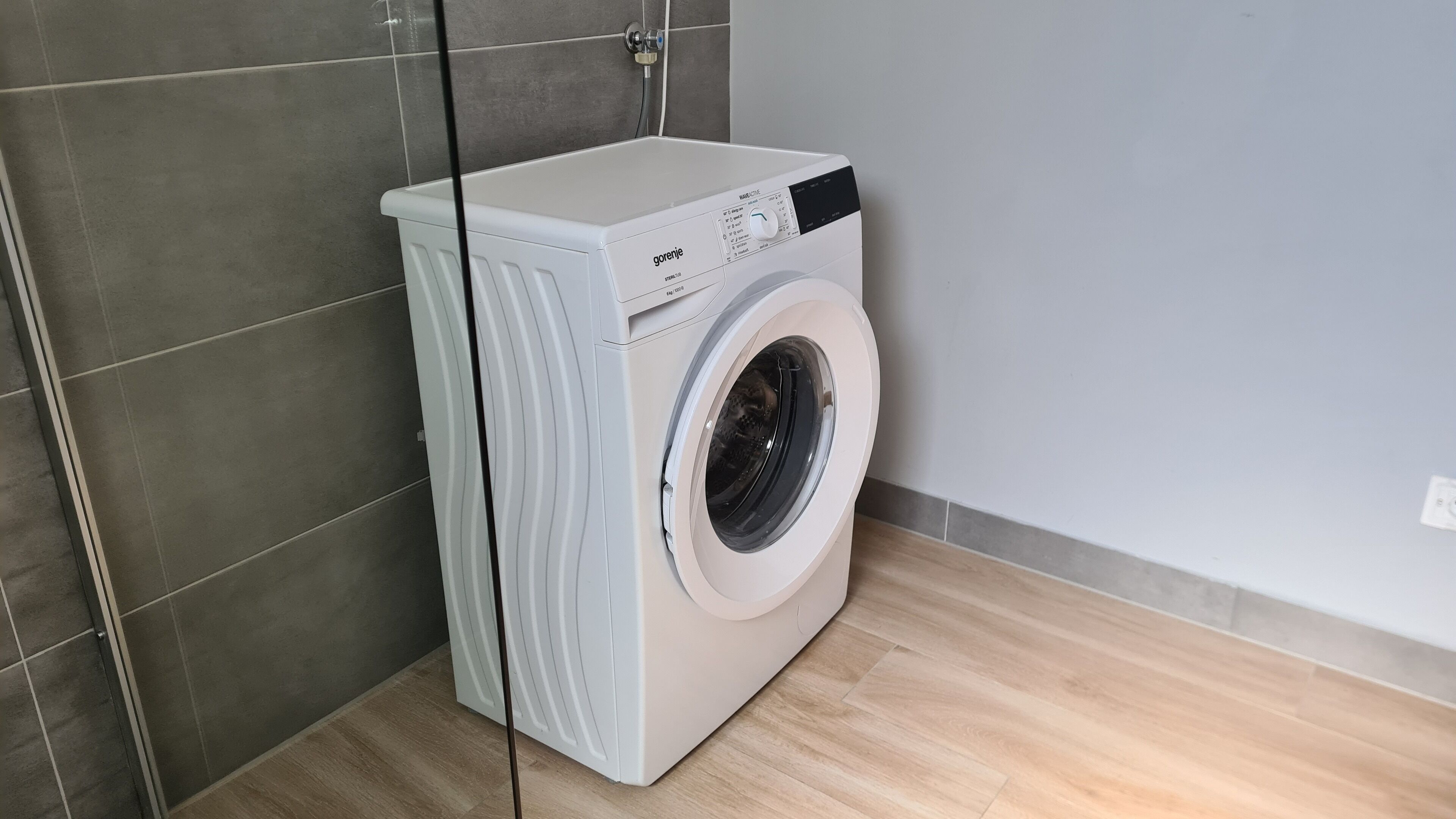 A compact, white front-loading washing machine in a modern bathroom with grey tiles and a wooden floor.