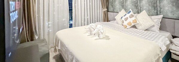Ένα καλοστρωμένο κρεβάτι με λευκά σεντόνια και διακοσμητικά μαξιλάρια, ένα γκρι προσκέφαλο και λεπτά, διάφανα κουρτίνια.